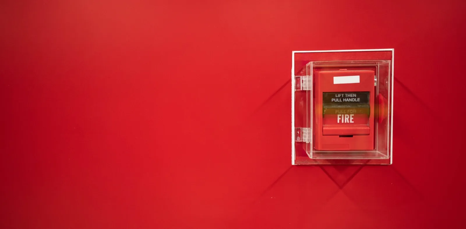 A dual-action fire alarm pull station under a clear protective cover, labeled "lift then pull handle," mounted on a red wall